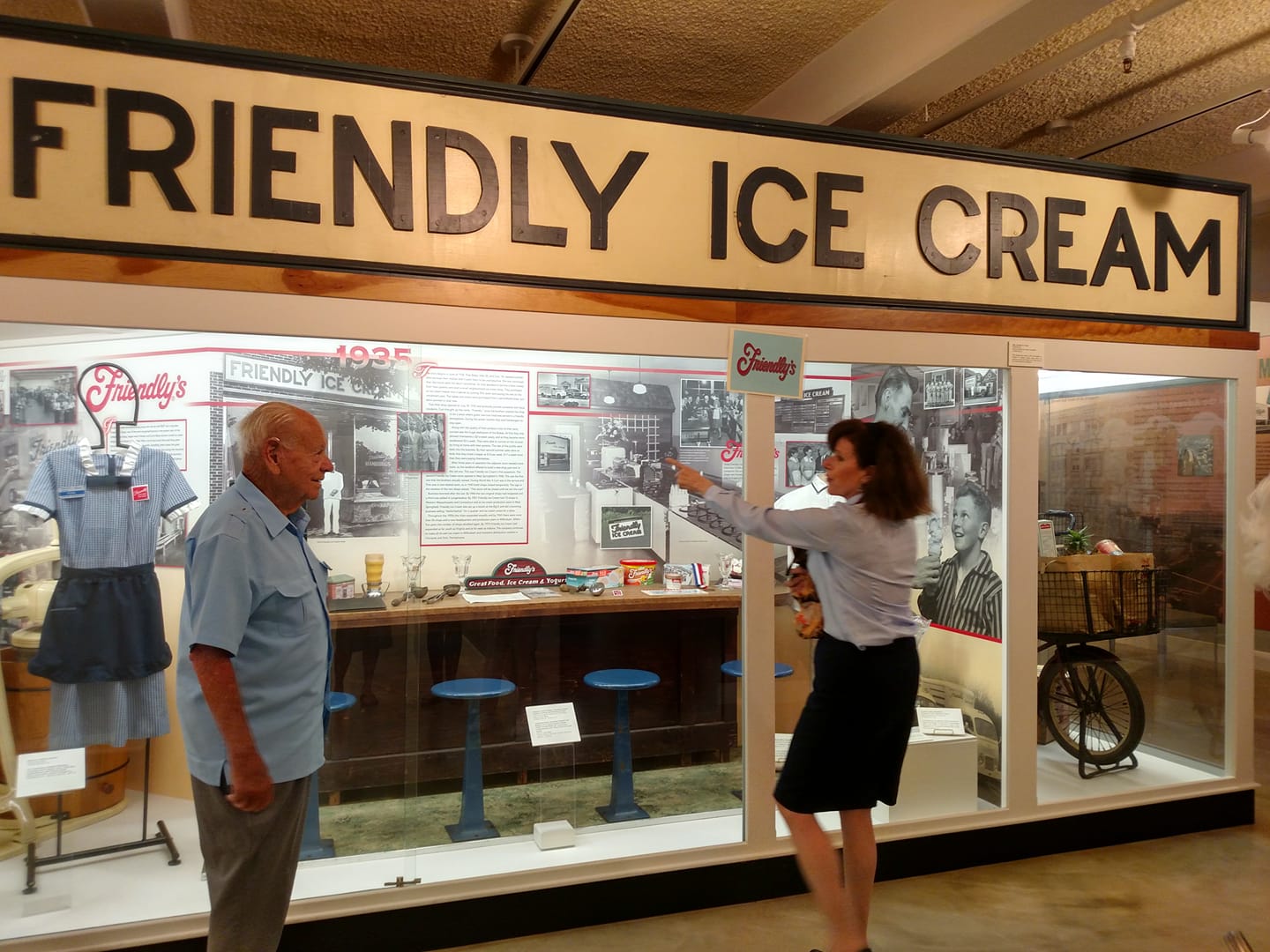 Old man stands in front of exhibit of Friendly Ice Cream while woman points to objects in the exhibit