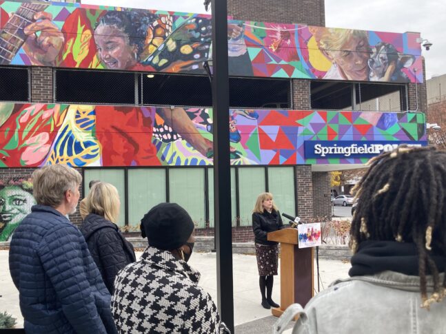 Crowd of people watching woman at podium speak outdoors under mural of children playing
