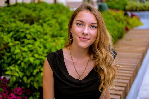 Photographic portrait of a smiling young white woman with long blond hair in front of a garden