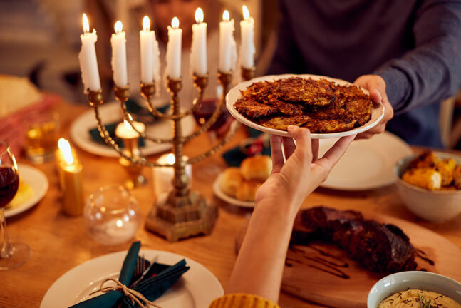 People passing latkes during a meal at dining table with a menorah at left.