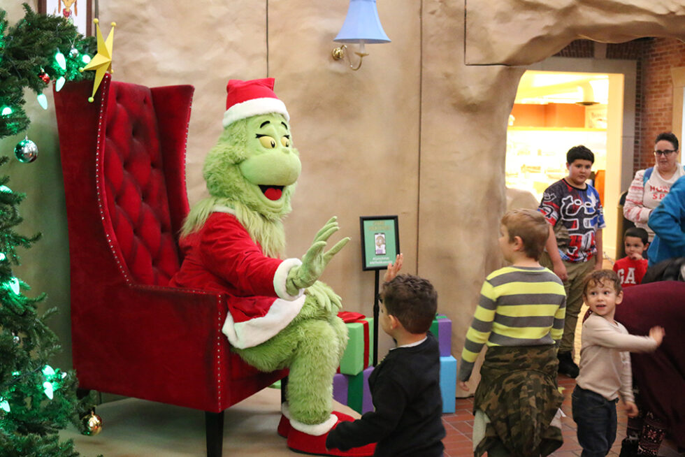 Dr. Seuss character the Grinch dressed as Santa Claus. He seated in a large chair, waving at children.