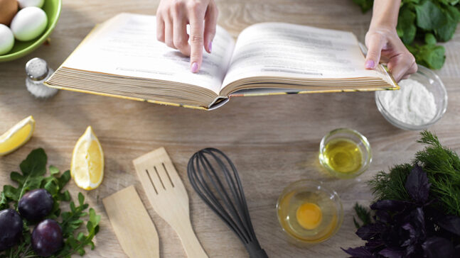 Woman reading recipe in a cookbook with kitchenware on table.