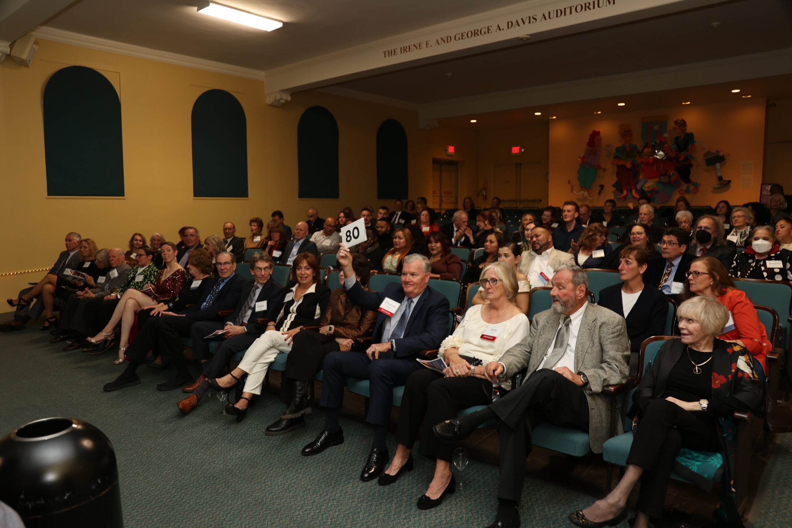 Crowd of people in an auditorium with one member of the crowd holding up their auction number