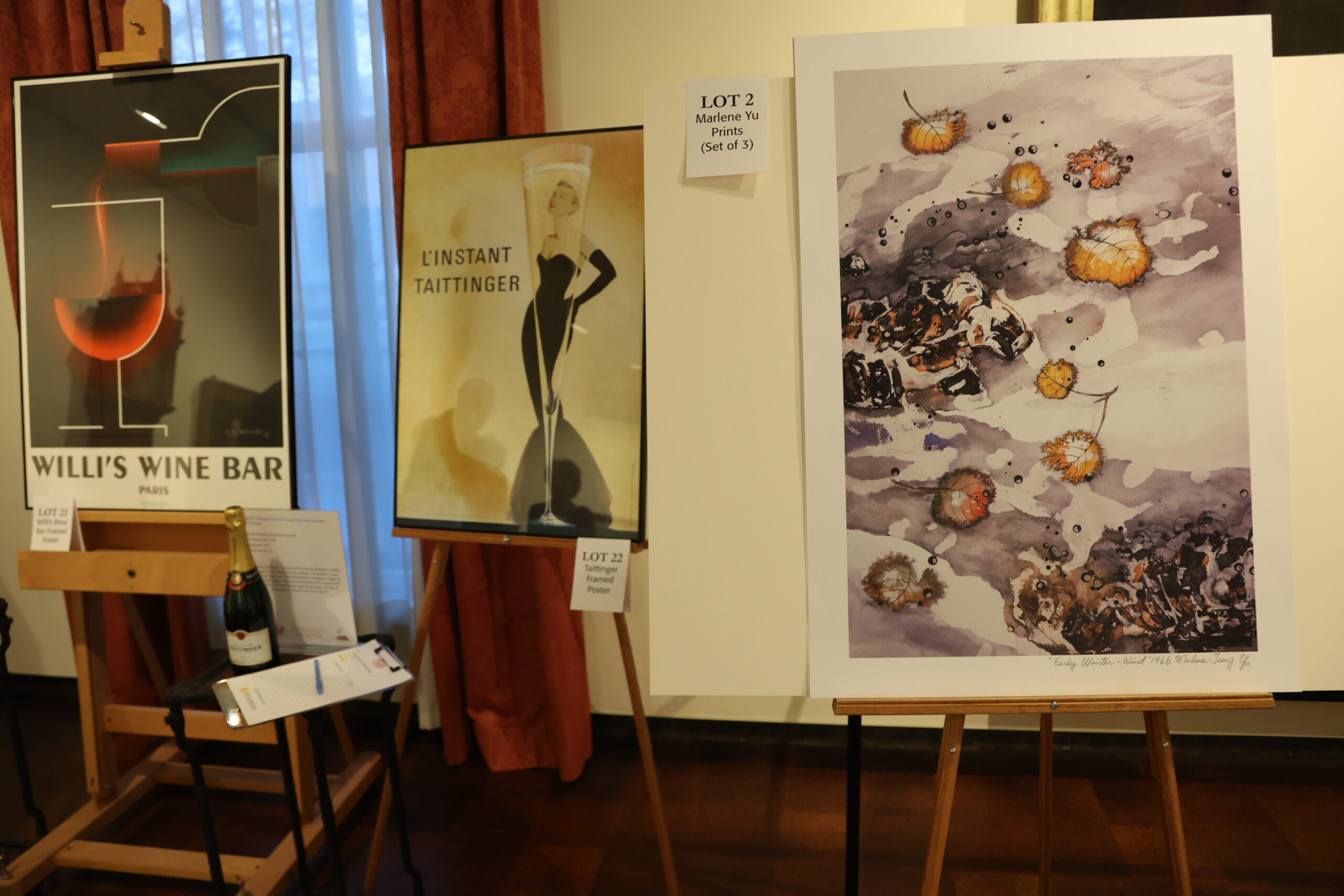 Three art pieces on easels and a wine bottle on a stand with a clipboard in front of it, each item is labeled with their auction lot number