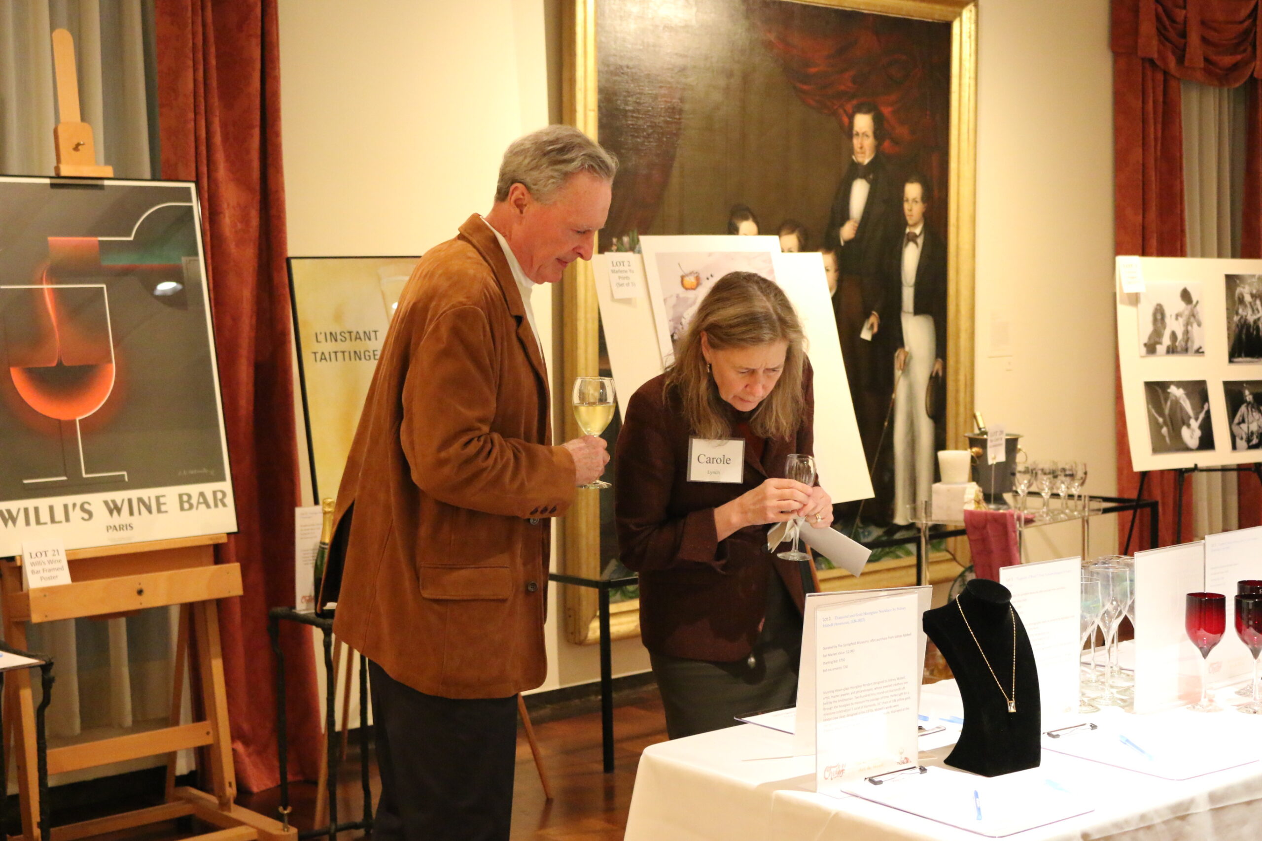 Two people holding wine glasses and looking at silent auction items on tables in a DMFA exhibit hall