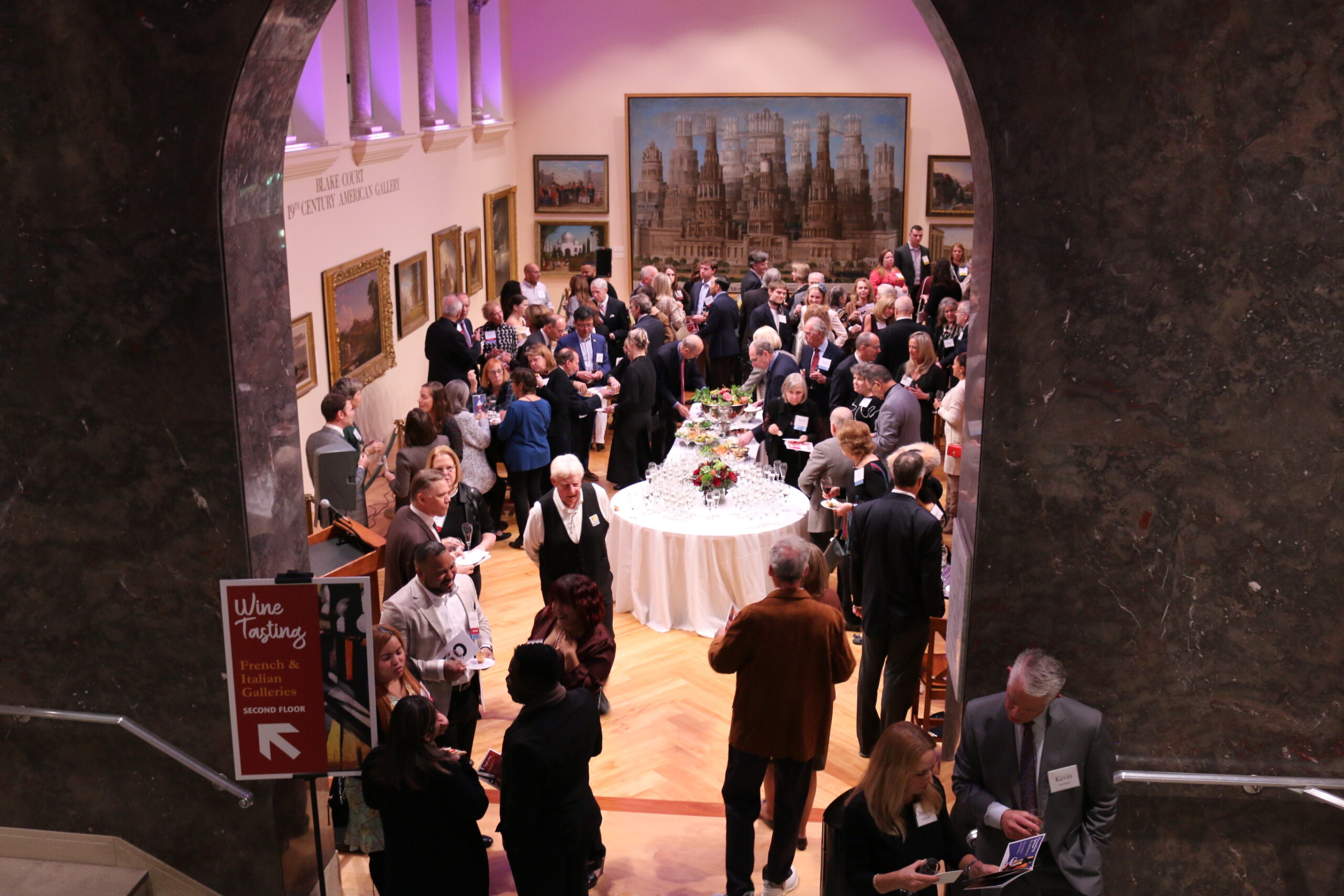 Wide shot of a crowd of people in the Blake Court art hall grabbing food and drinks or conversing, as seen through the arch of the Blake Court entrance