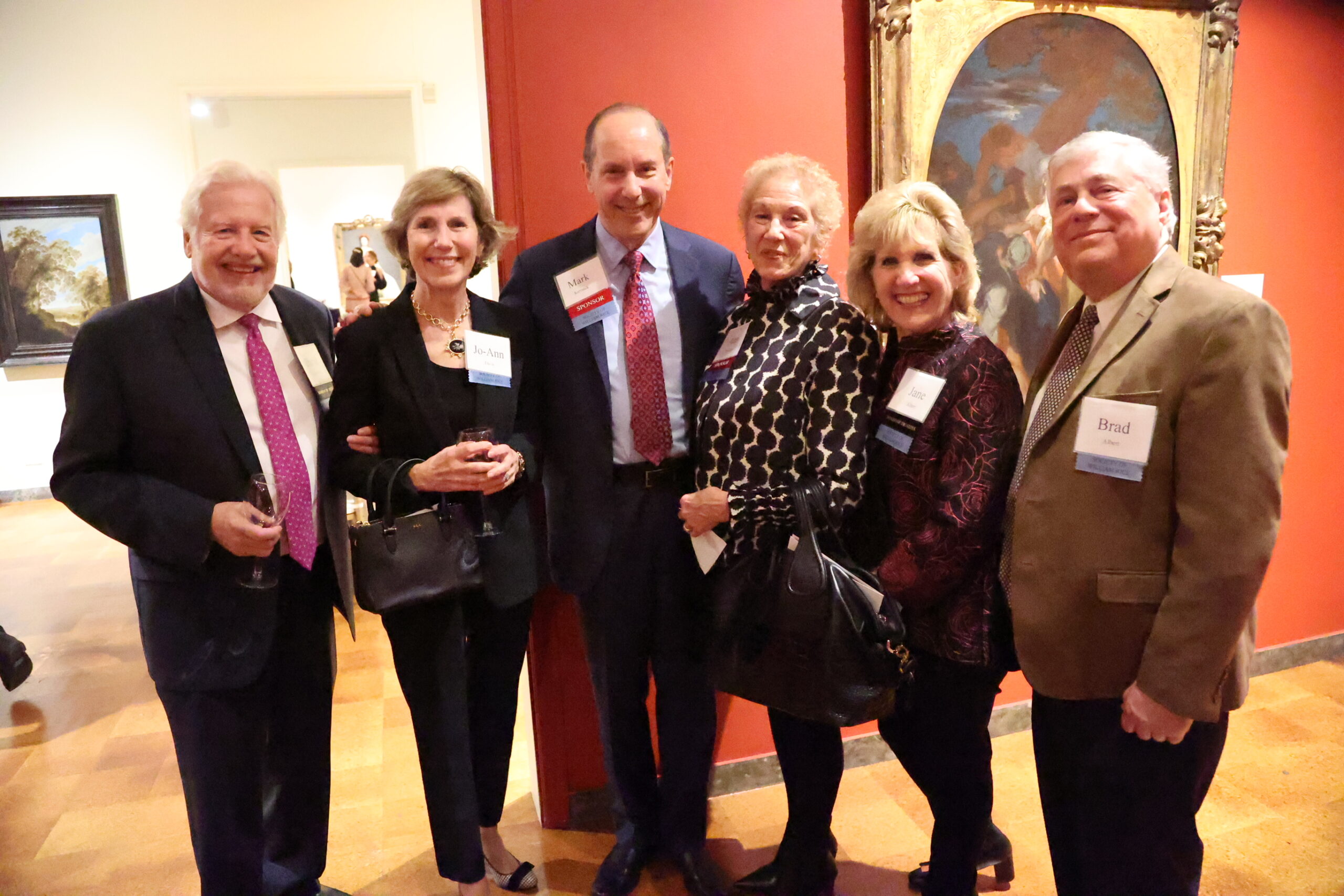 Six people lined up together and smiling for the camera, two of whom are holding a wine glass