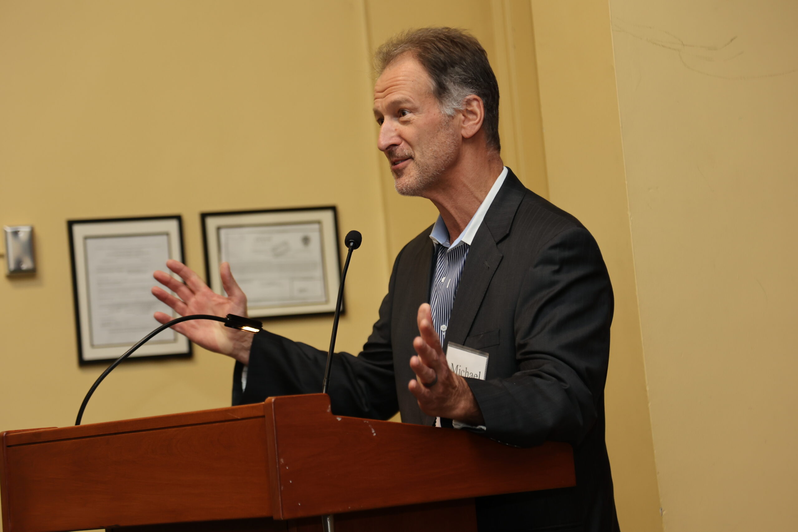 Mike Matty standing and speaking at a podium, in the midst of gesturing with his hands