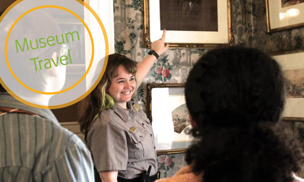 Smiling ranger points to artwork on the wall, while two visitors facing her look on A guided tour in the Longfellow House parlor.
