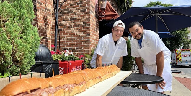 Two men posing with a long sub sandwich