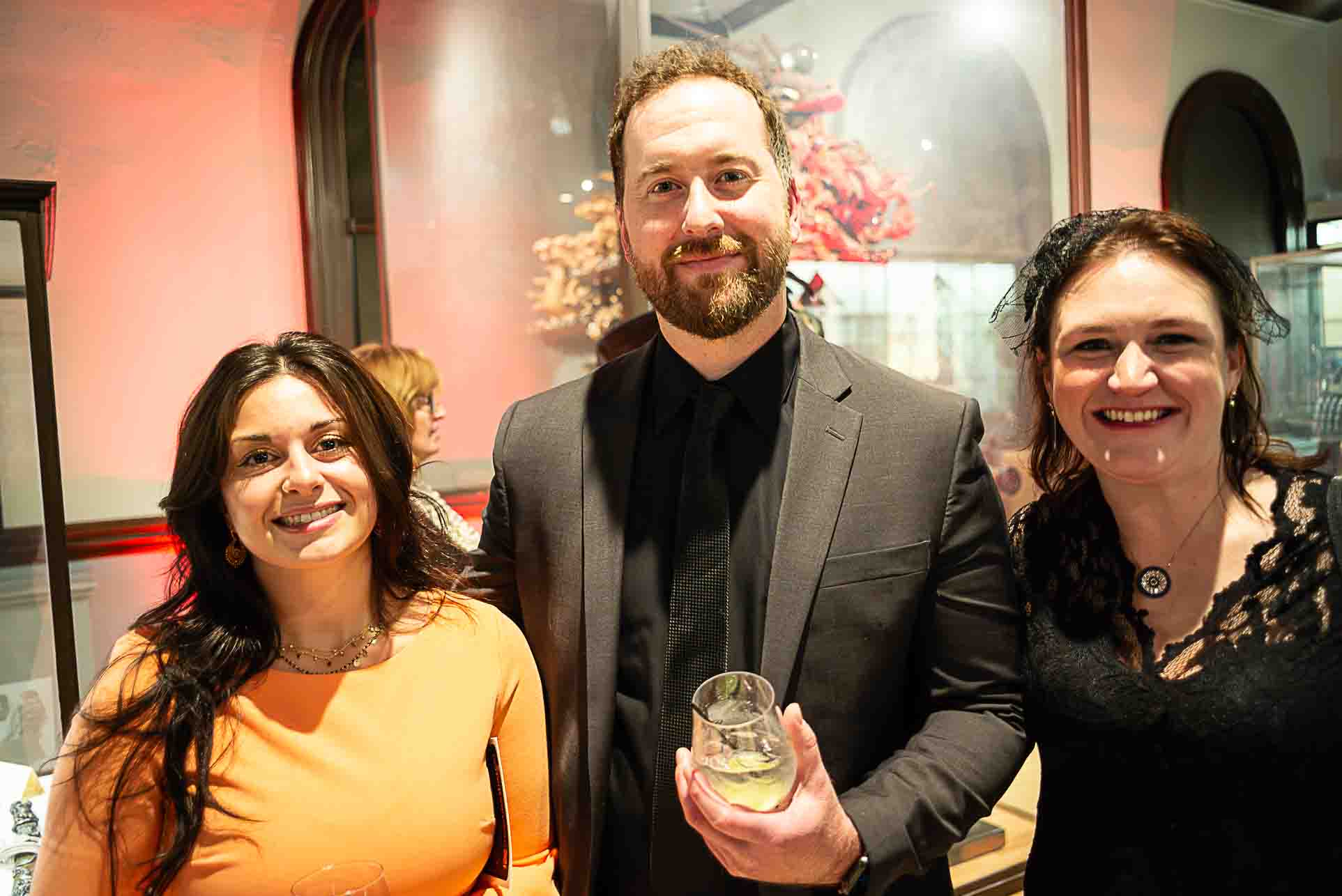 Three people smiling at a museum event, standing close together with drinks in hand.