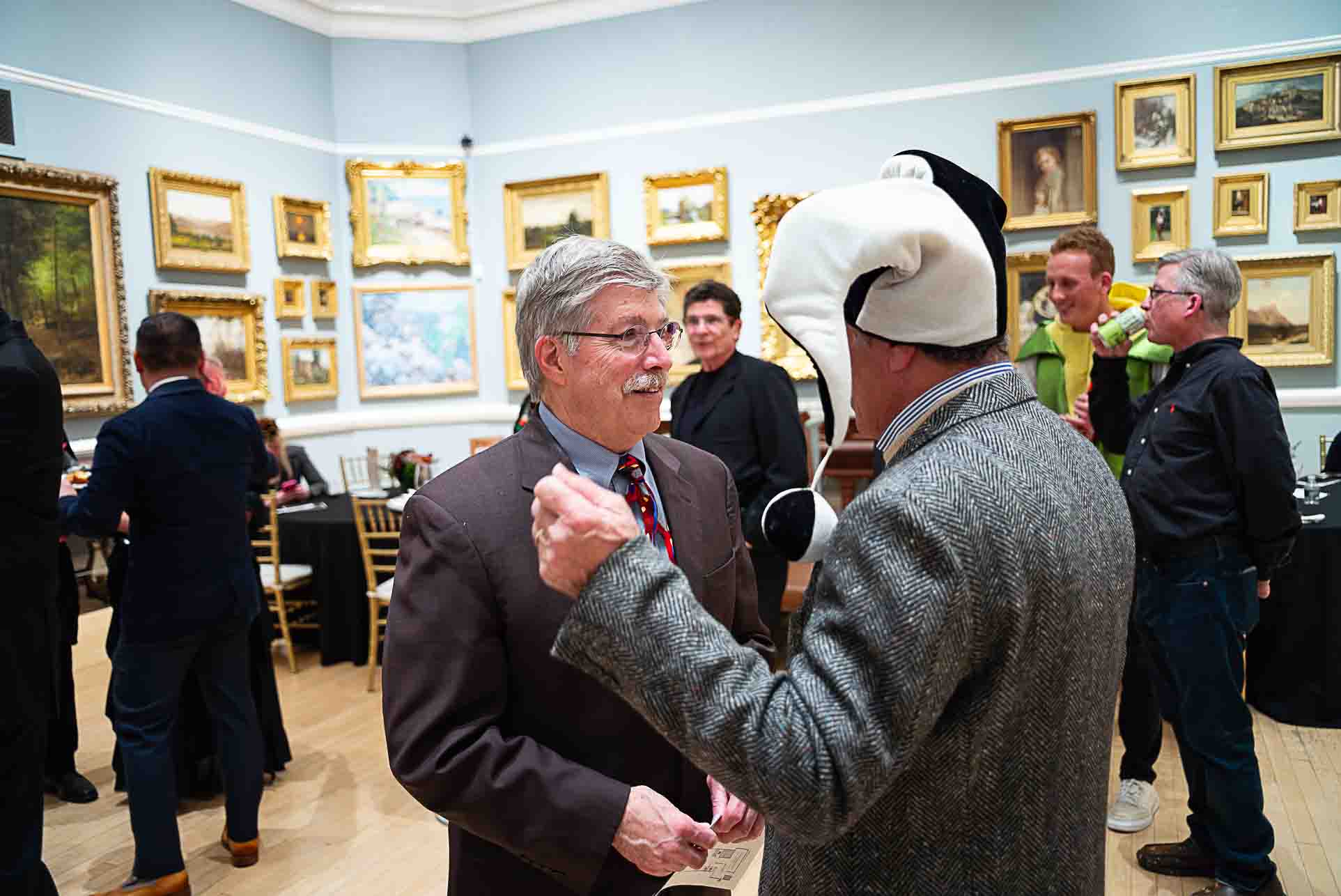 Guests chatting in an art gallery, including one person wearing a playful black-and-white costume hat.