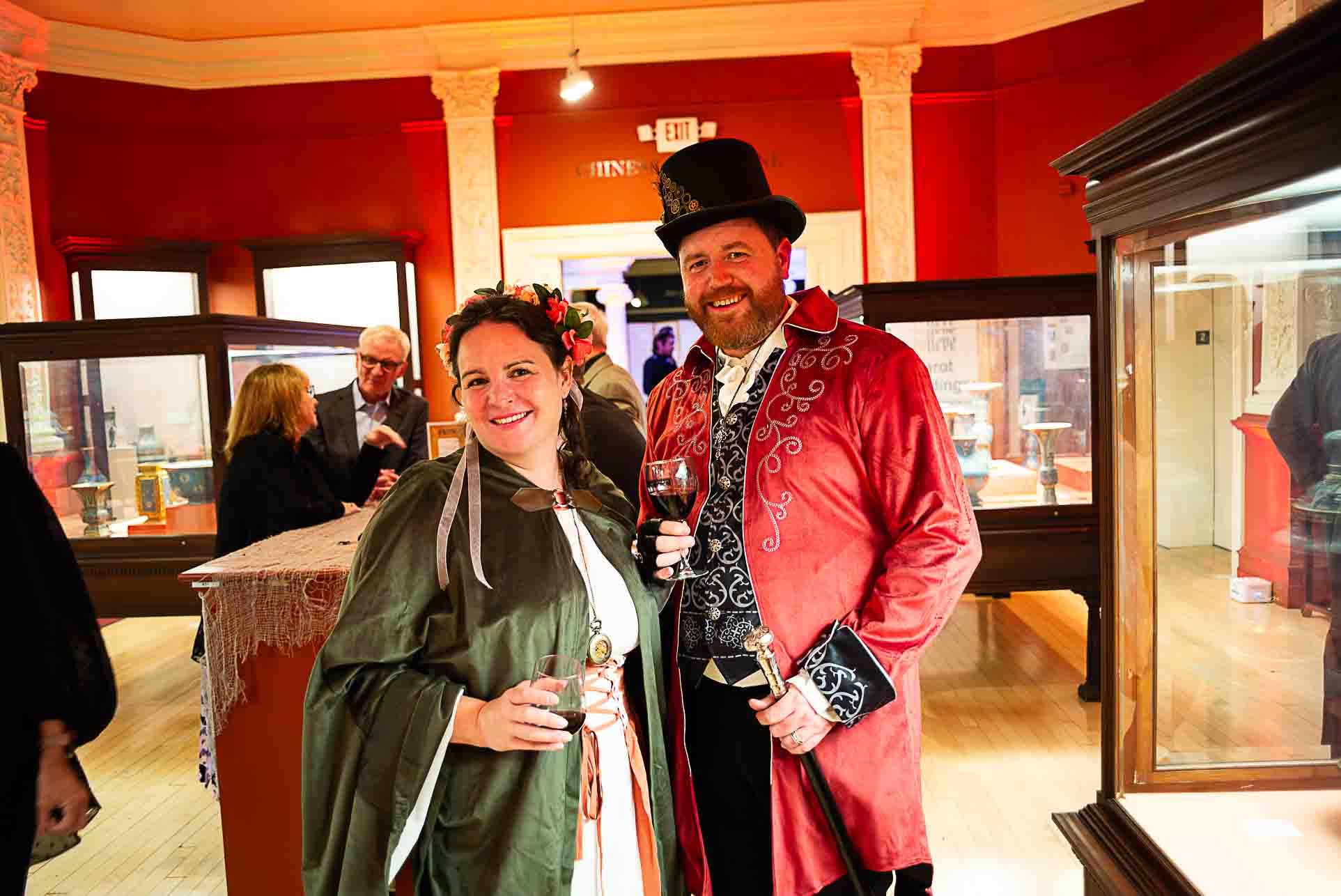 Two guests in festive, vintage-style costumes smile and hold drinks in a museum gallery.
