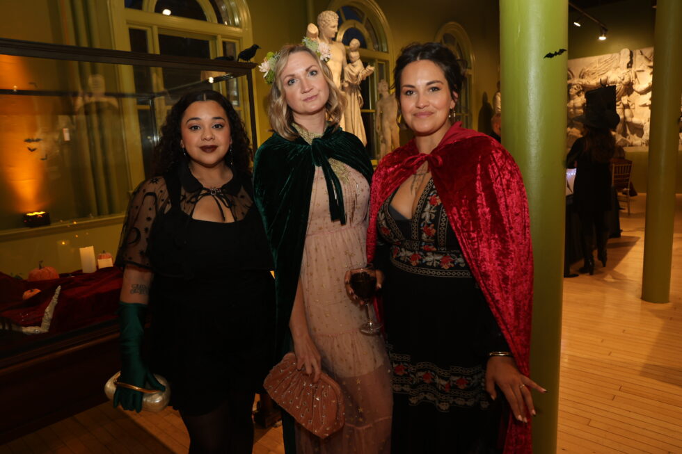 Three women dressed in semi-formal attire with capes and accessories stand together at a museum event with festive decor and warm lighting.